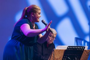Eine blonde Frau singt vor blauem Hintergrund in ein Mikrofon, sie gestikuliert mit der rechten Hand. Foto: Mareen Meyer/Grimme-Institut