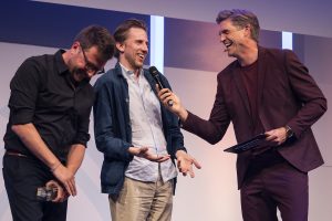 Drei weiße Männer stehen lachend auf einer Bühne, der rechte hält ein Mikrofon. Foto: Arkadiusz Goniwiecha/Grimme-Institut